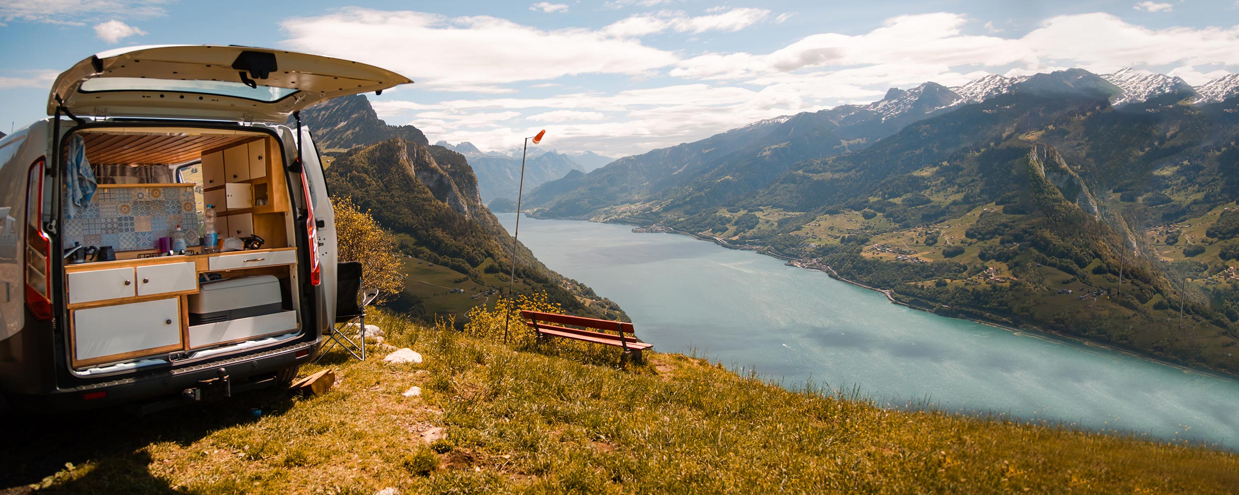 Van conversion with kitchen setup on a mountain top overlooking a lake and mountains.